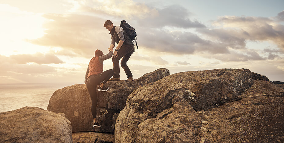 Photo of a male hiker helping a female hiker climb onto a boulder, with a sunset over the beach in the background